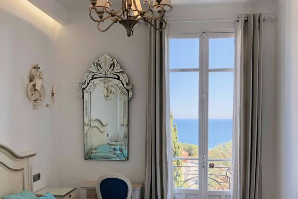 A bedroom with a bed, a desk, a chair, a chandelier, a large mirror, and a view of the sea.