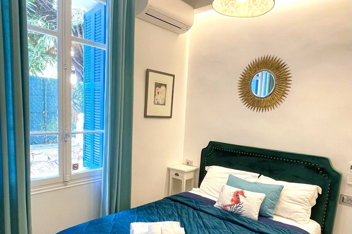 A neatly made bed with a blue bedspread and pillows, a small bedside table with a lamp, a wall-mounted mirror, and a window with curtains.