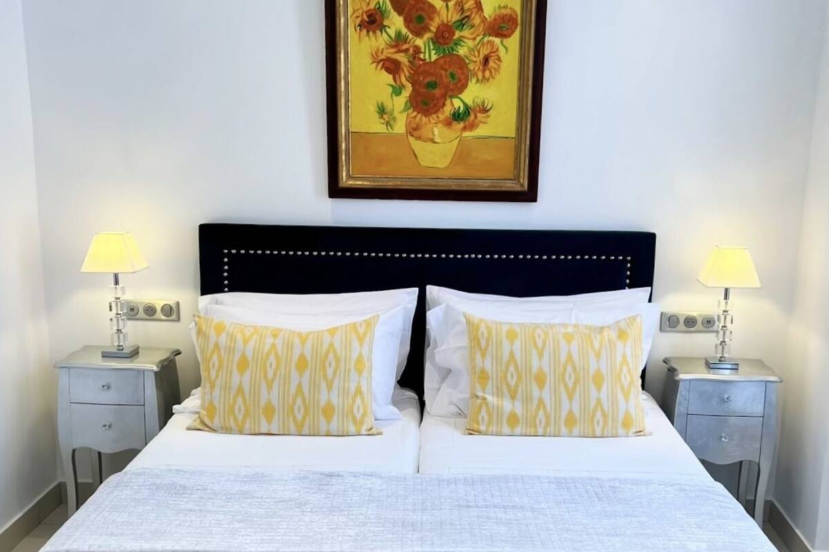 A neatly made double bed with a quilted white bedspread, two matching pillows with a yellow and white pattern, and a framed painting of sunflowers on the wall above.