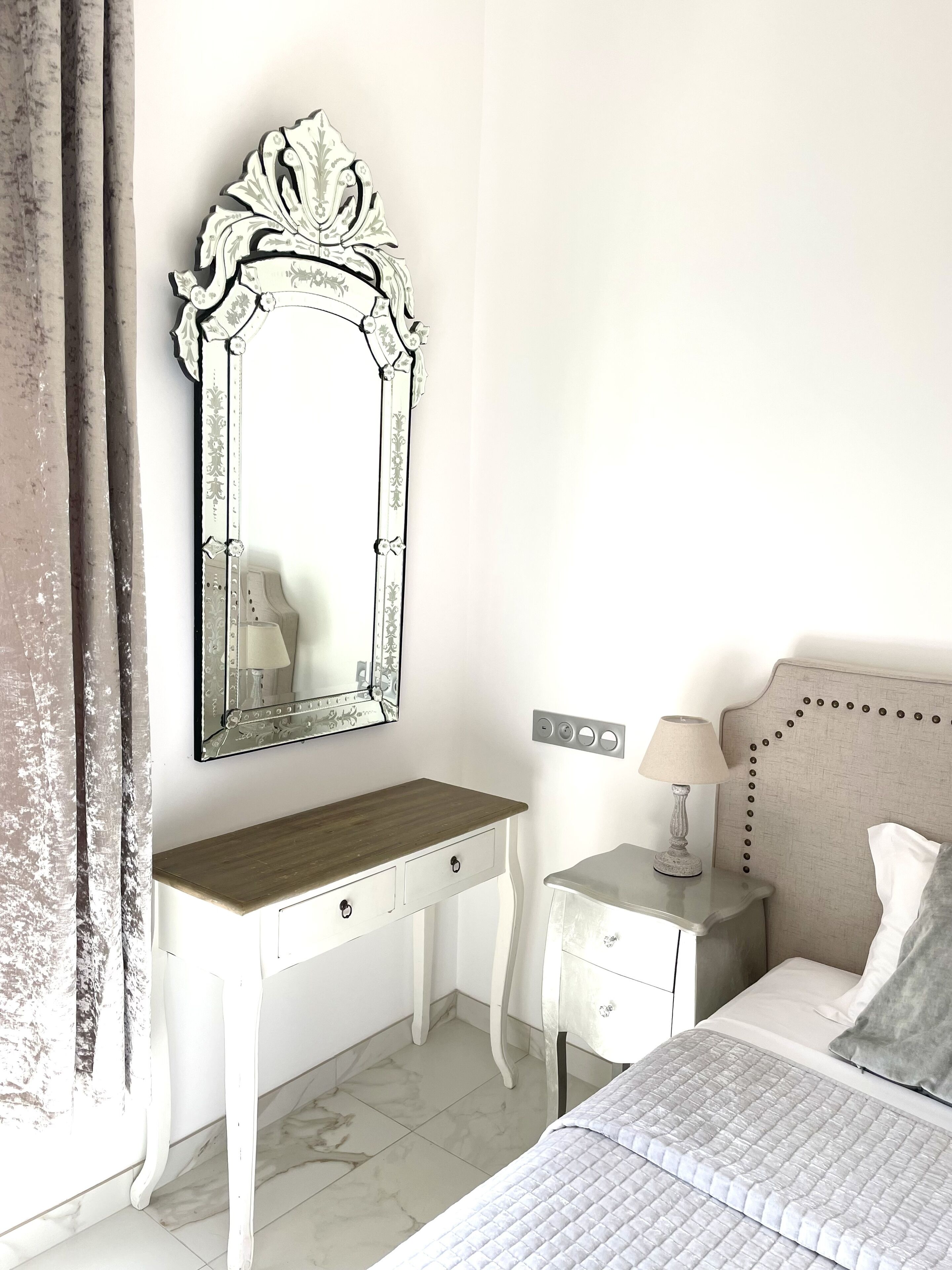 A bedroom with a white vanity, a large ornate mirror, a bedside table with a lamp, and a bed with a quilted cover.