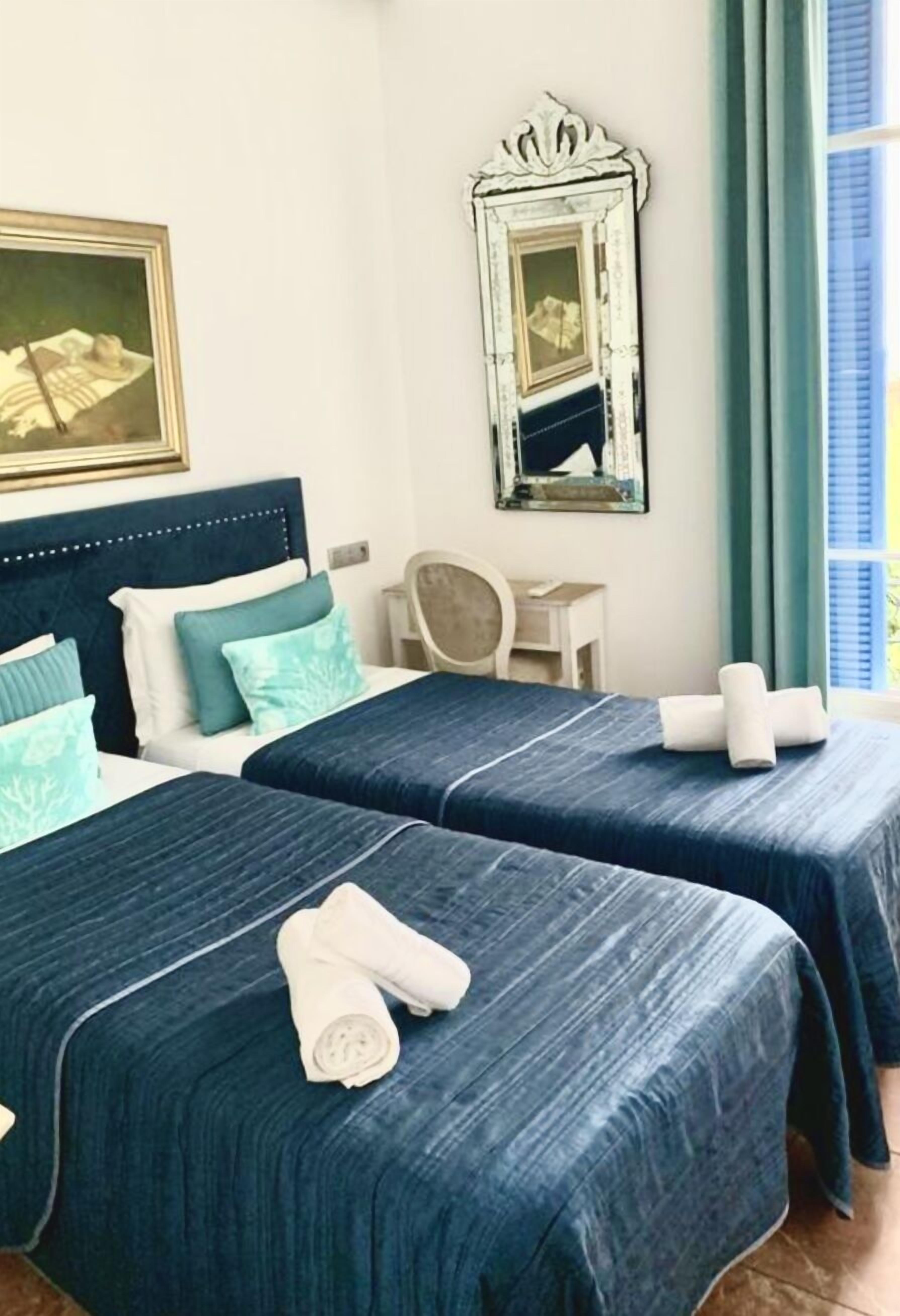 Et hotelværelse med to senge, hver med et blåt sengetæppe og hvide puder. Der er et spejl på væggen og en stol ved siden af sengen.