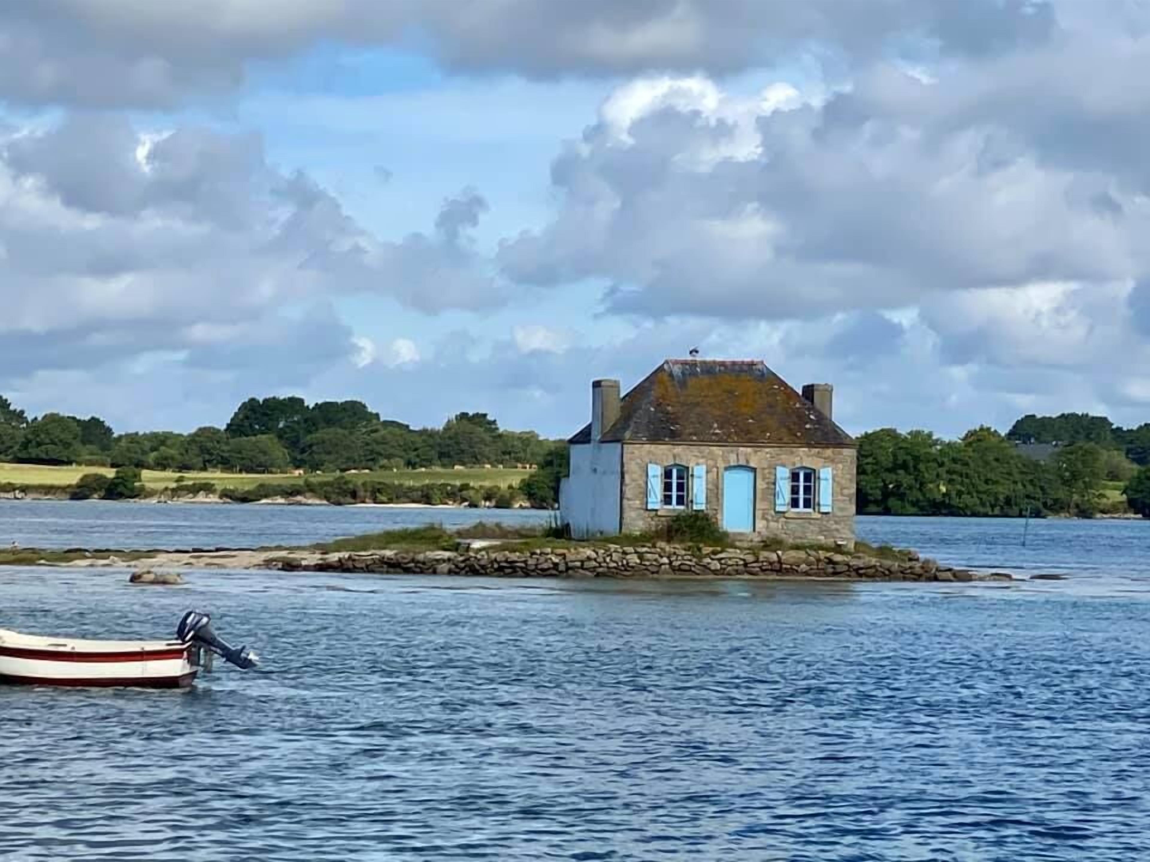 Ein kleines Steinhaus mit blauen Fensterläden auf einer felsigen Insel, umgeben von Wasser.