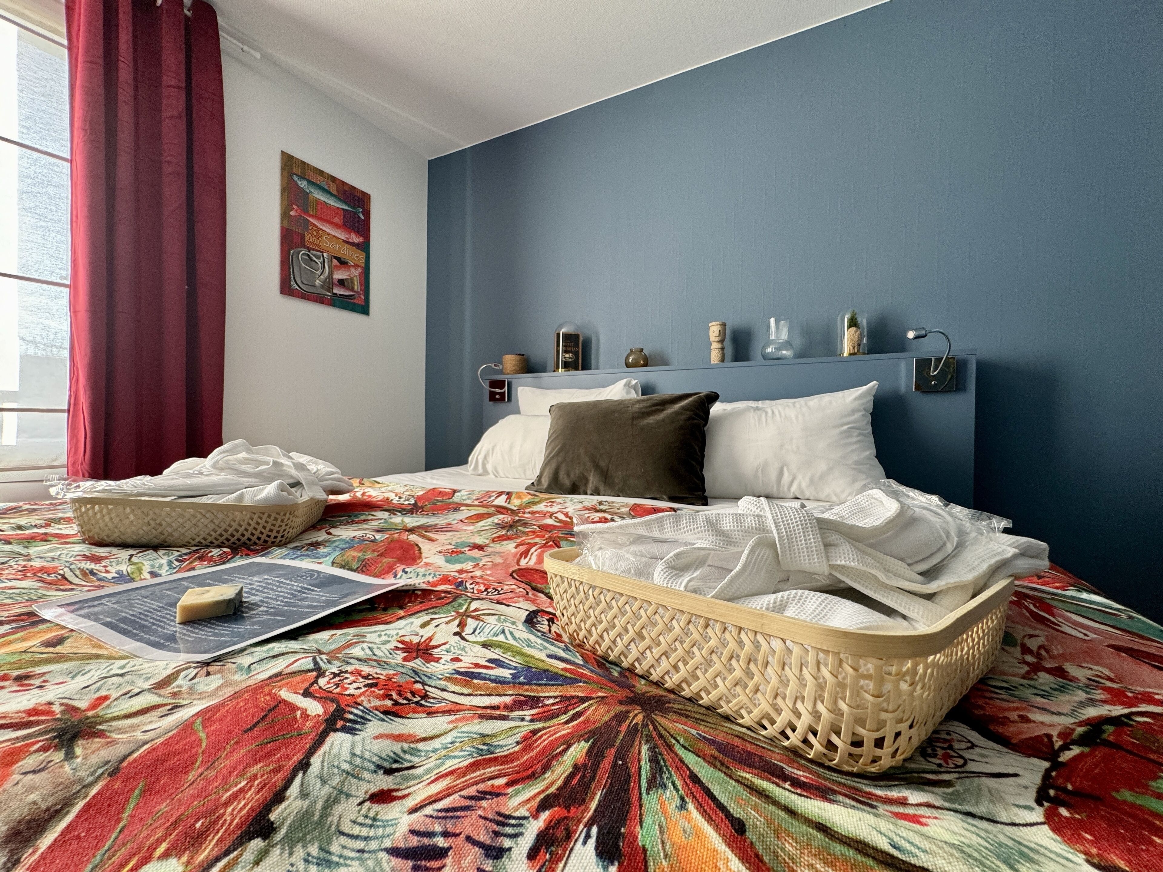 A bedroom with a bed, a headboard with shelves, a window with red curtains, and a wall with a framed picture.