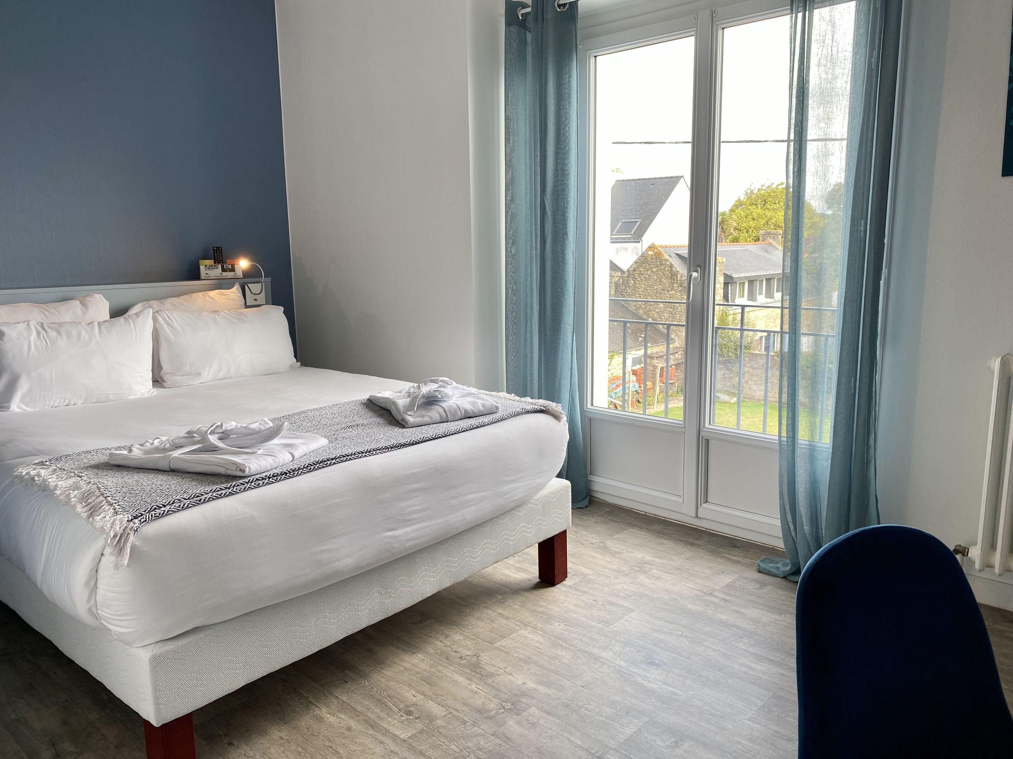 Una cama bien hecha con una colcha estampada, una mesita de noche con una lámpara y vistas a una casa a través de un ventanal con cortinas azules.