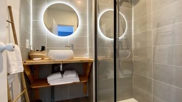 Un baño moderno con ducha envuelta en vidrio, tocador de madera con lavamanos y dos espejos iluminados.
