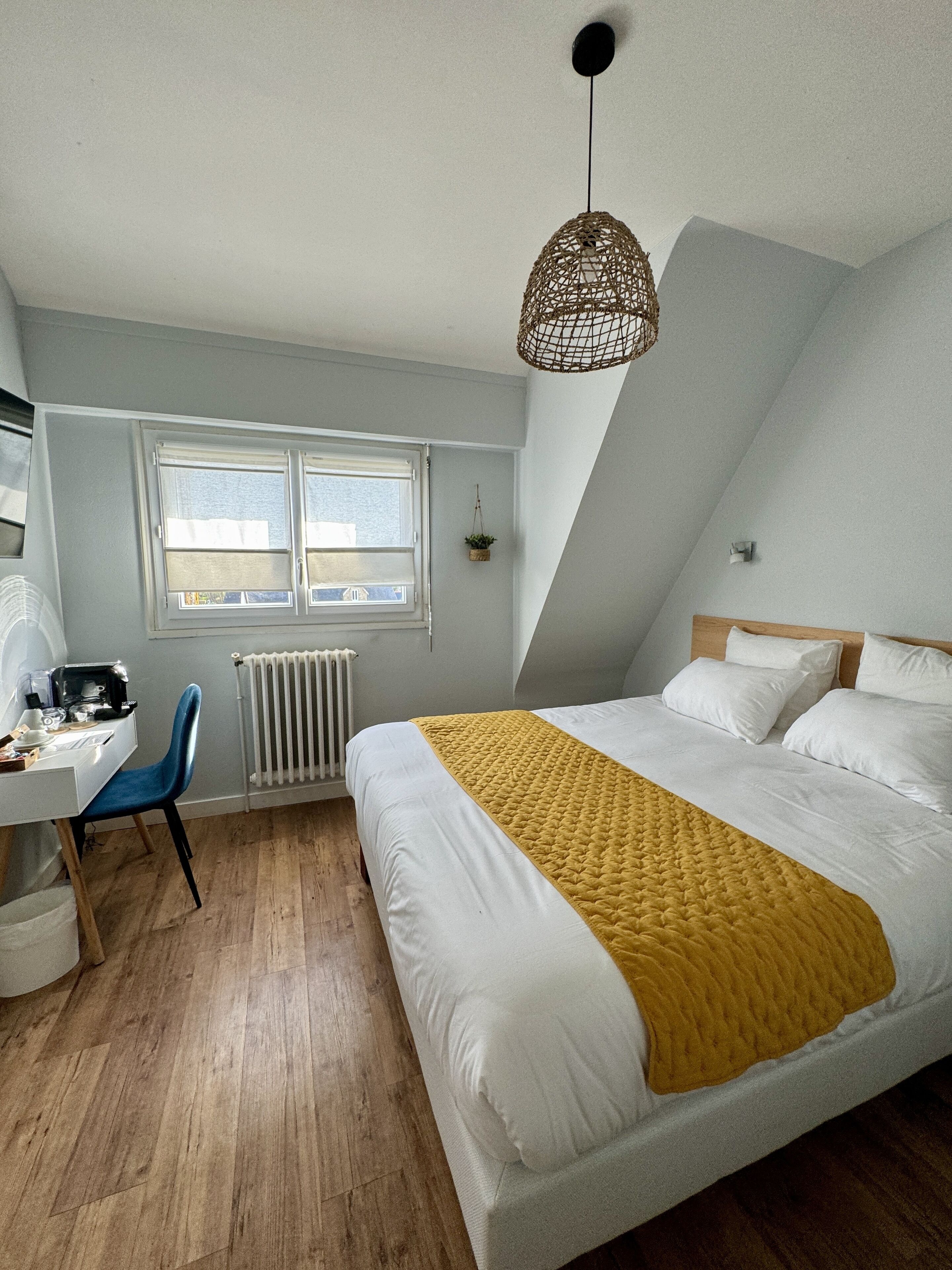 A bedroom with a bed, a desk, a chair, a window, and a hanging light fixture.