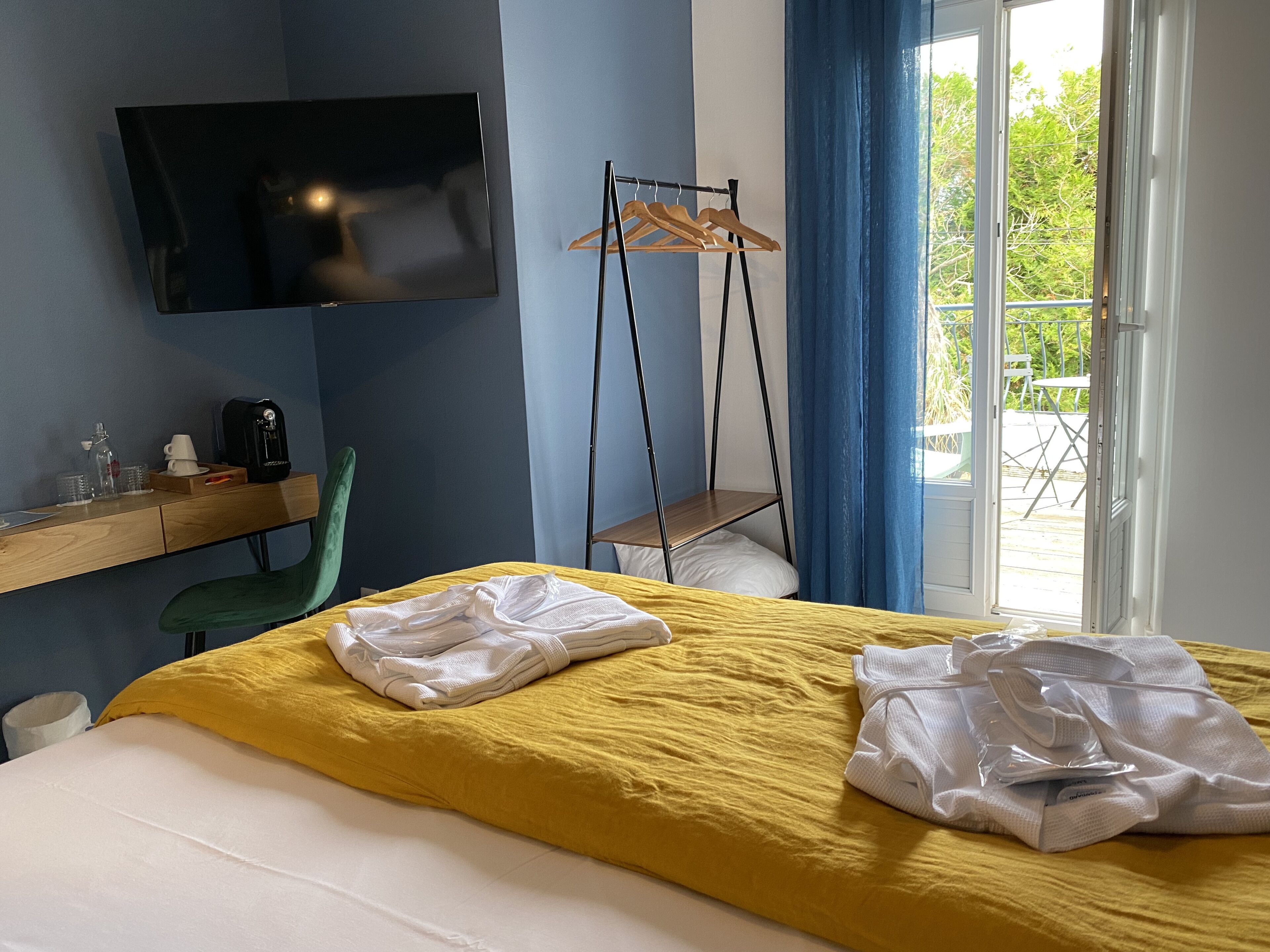 A hotel room with a bed, a TV mounted on the wall, a desk with a chair, a hanging clothes rack, and a view of the outdoors through a door.