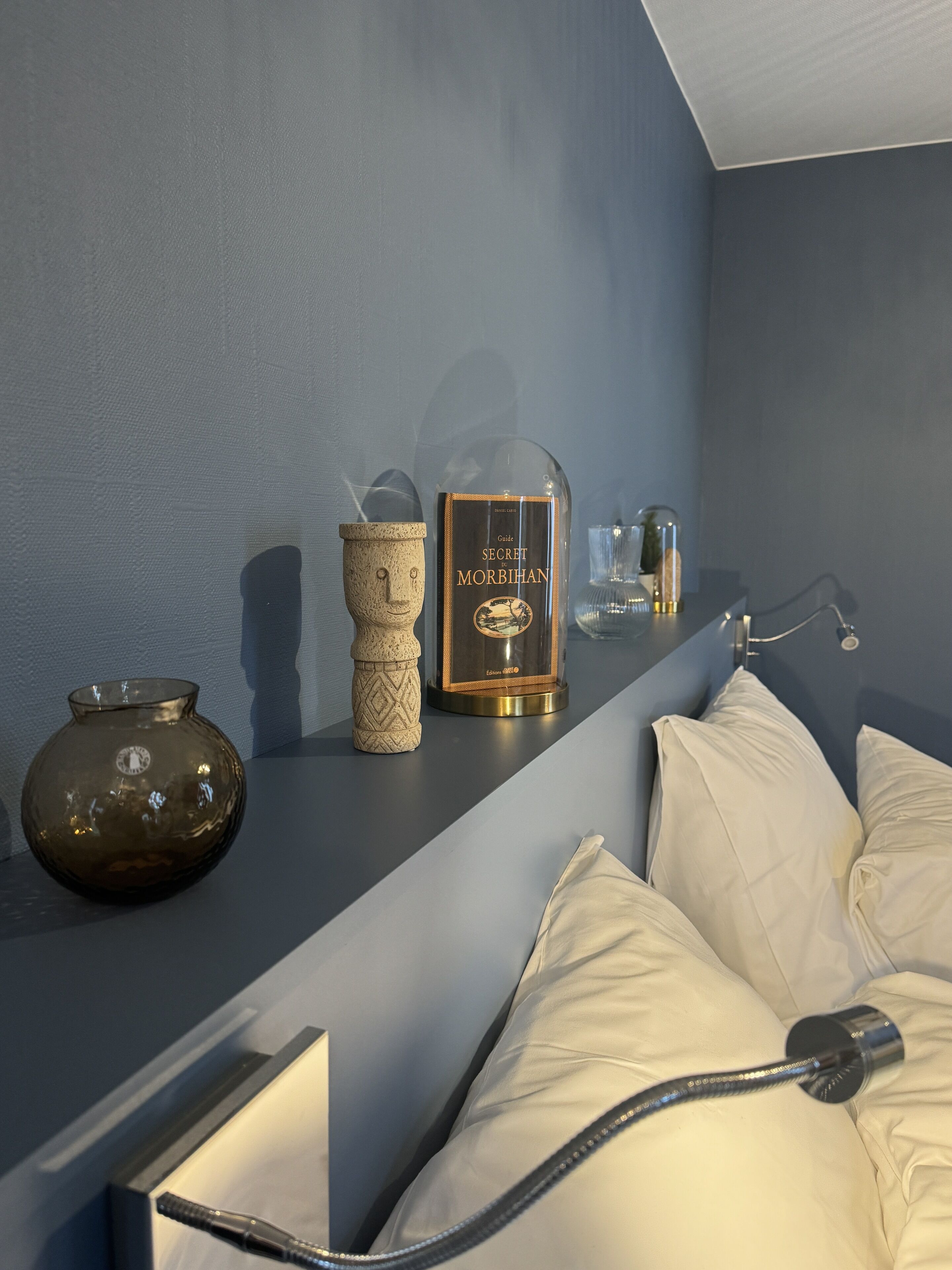 Une chambre à coucher avec un lit, des oreillers, une table de chevet avec un livre, un vase et une lampe.