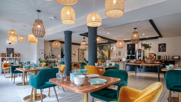 Un espace de restauration moderne avec des chaises turquoise et jaunes, des tables en bois et des suspensions lumineuses en fibres tressées.