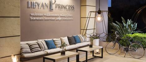 A modern lobby with a sofa, tables, a bicycle, and a wall with multilingual welcome text.