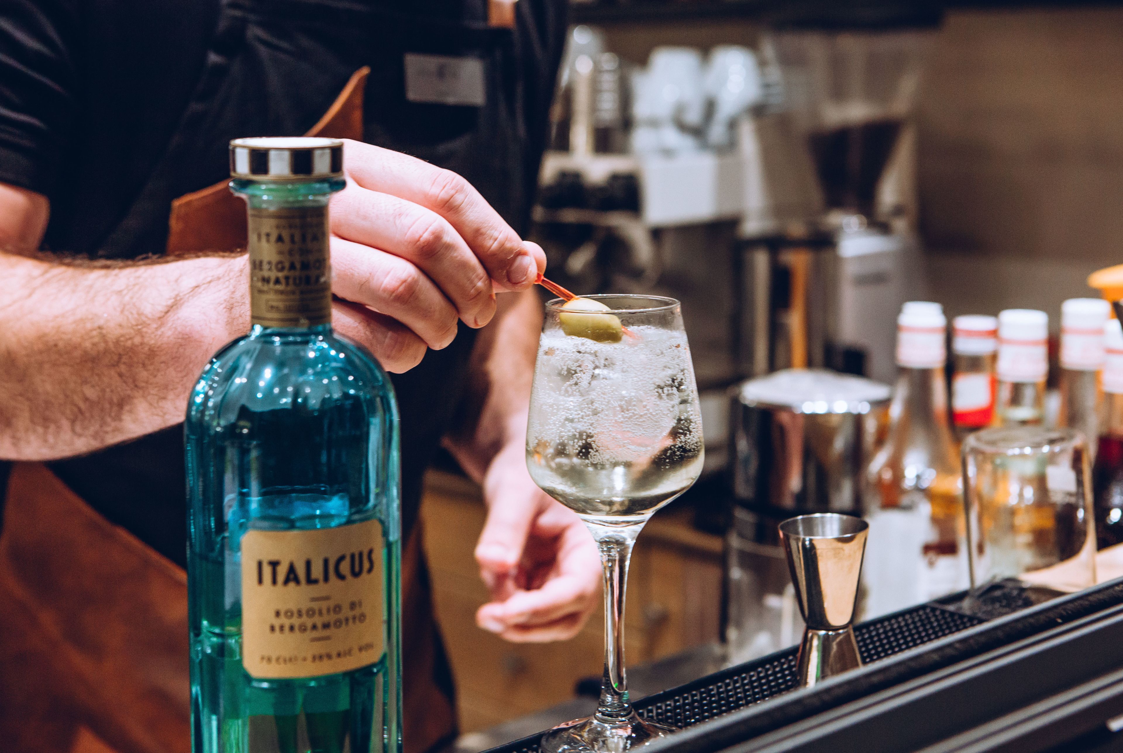 Ein Barkeeper bereitet ein Getränk zu, mit einer Flasche mit der Aufschrift "ITALICUS" sowie einem Glas mit Eiswürfeln und einer Limettenscheibe.