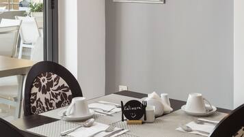 A dining area with a table set for two, a decorative light fixture, framed pictures on the wall, and a view into another room.