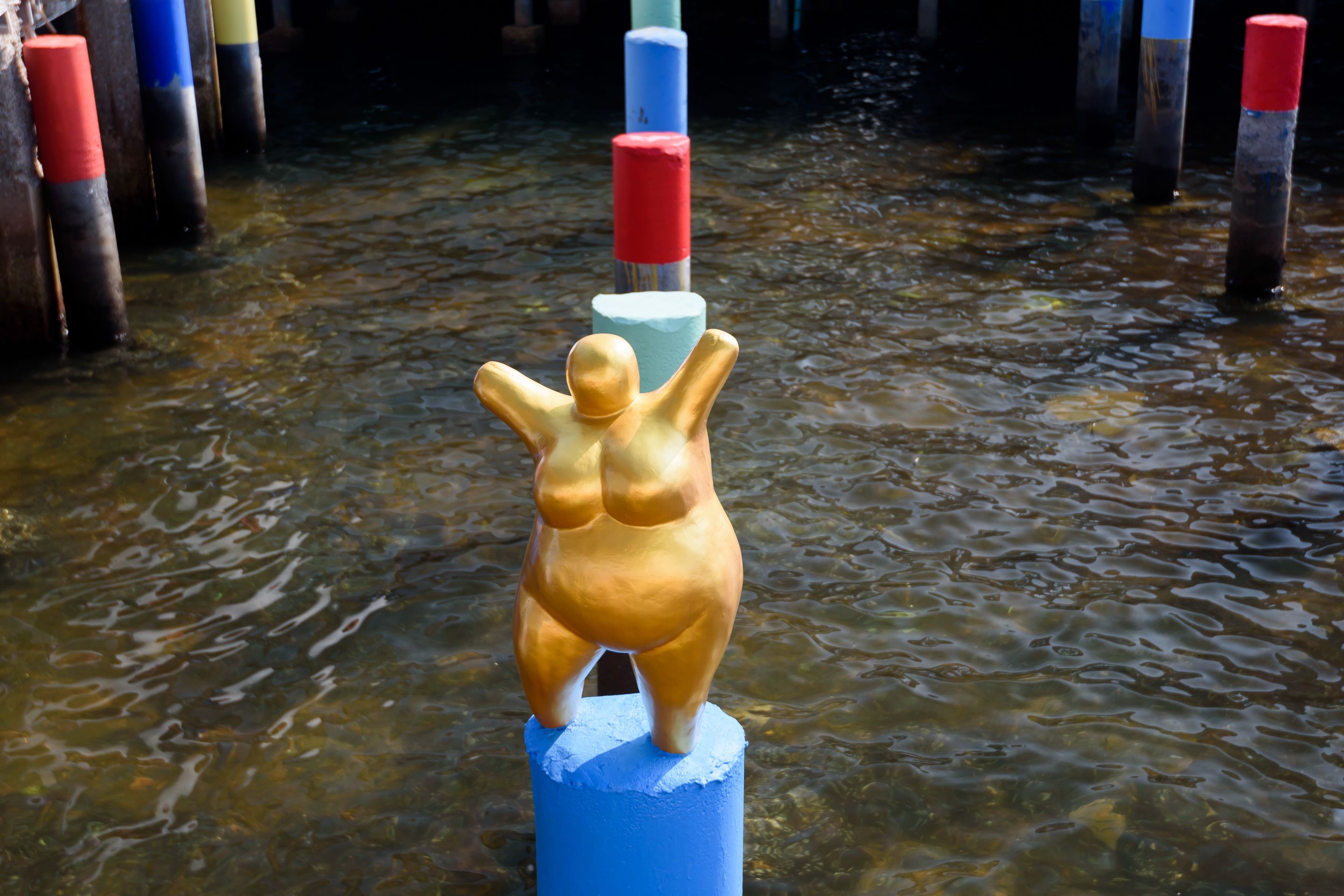A sculpture of a figure with raised arms on a blue post in a body of water.
