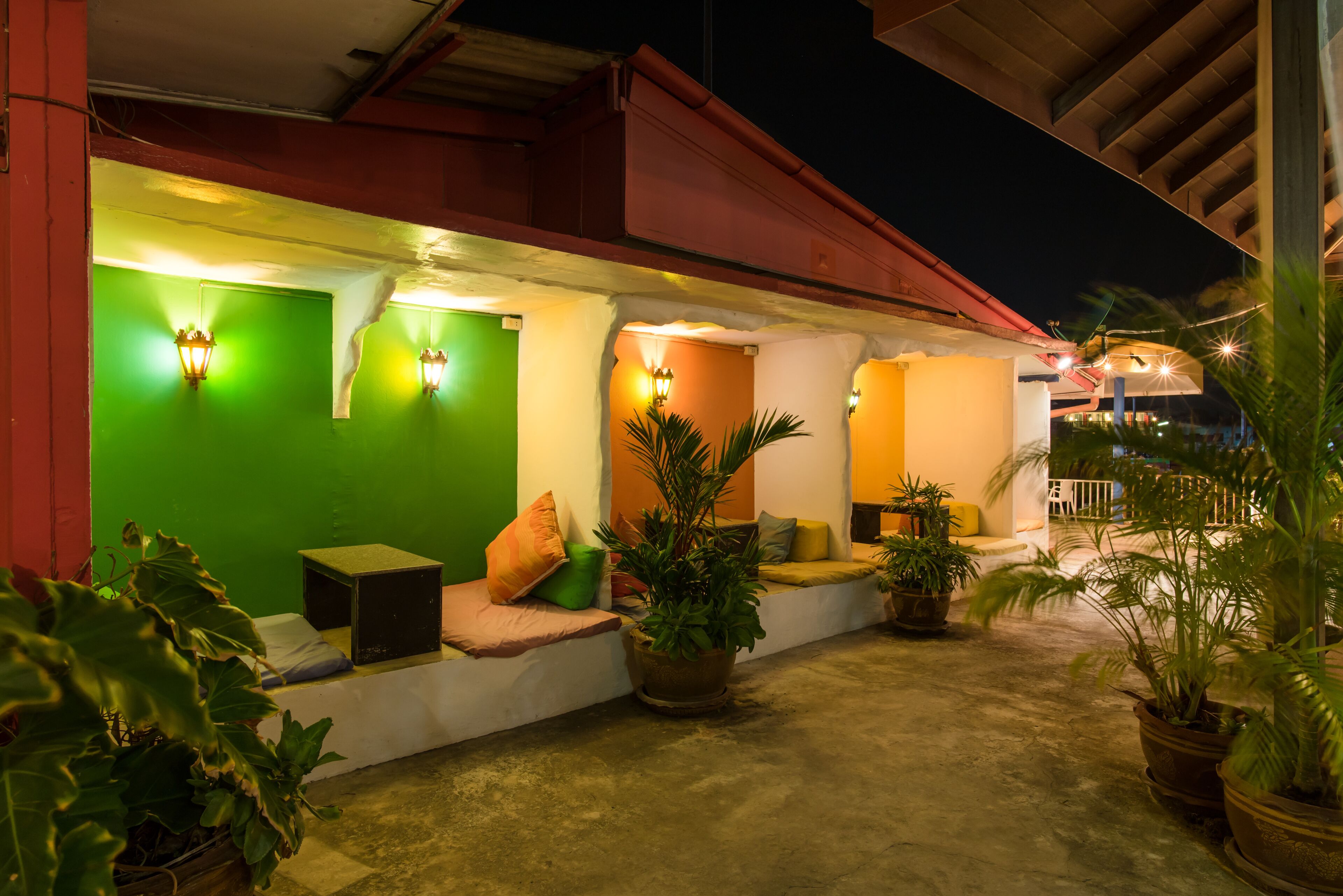 Un área de estar al aire libre con pared verde, pared naranja y pared blanca, cada una con luces fijadas en la pared. Hay varias plantas en macetas y una pequeña mesa con un altavoz.