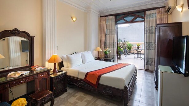 A hotel room with a large bed, a desk with a lamp, a television, and a balcony with outdoor furniture. - Lucky Hotel (Hanoi)