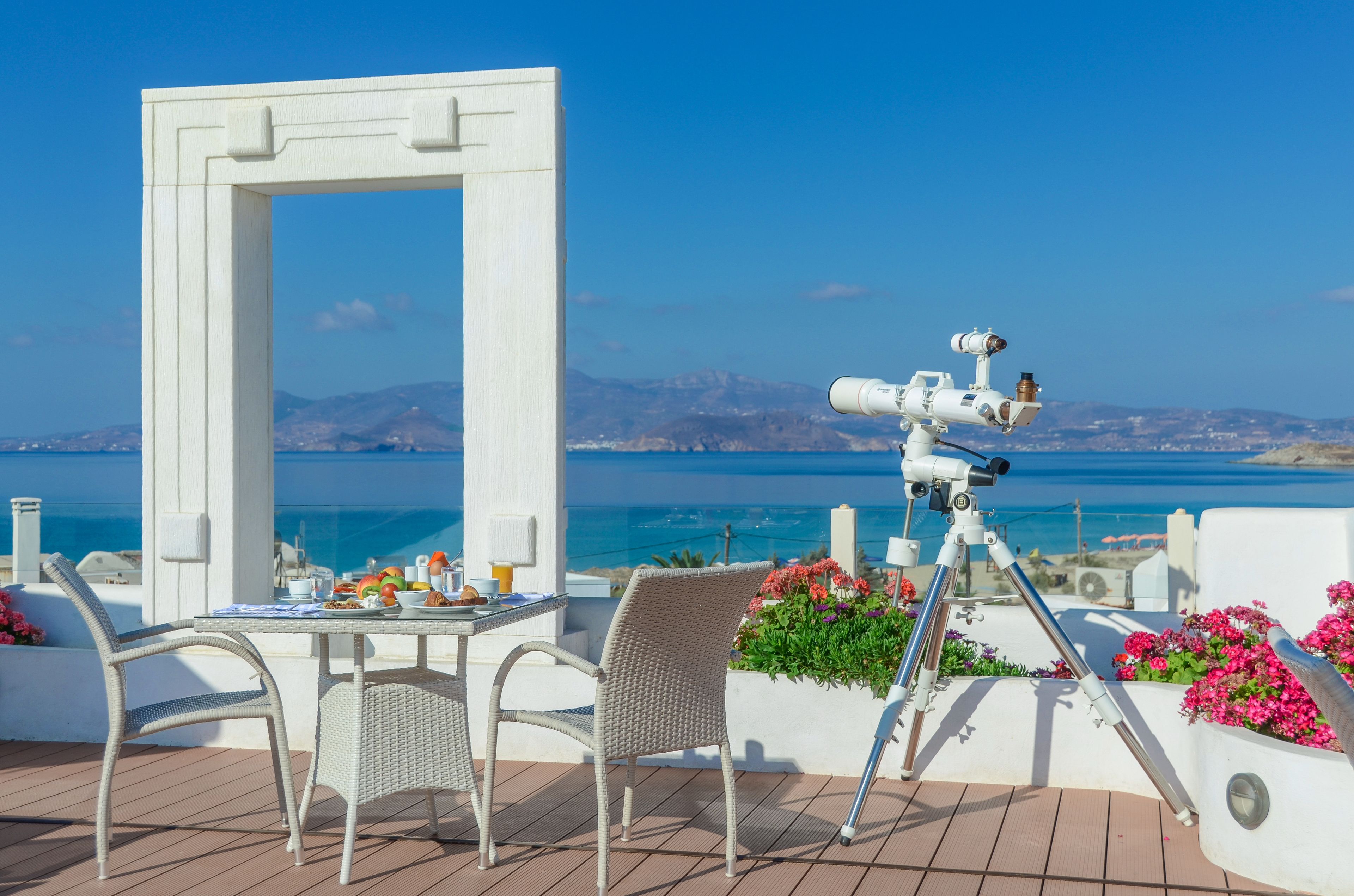 A terrace with a telescope, wicker chairs, a table with breakfast, and a view of the sea.