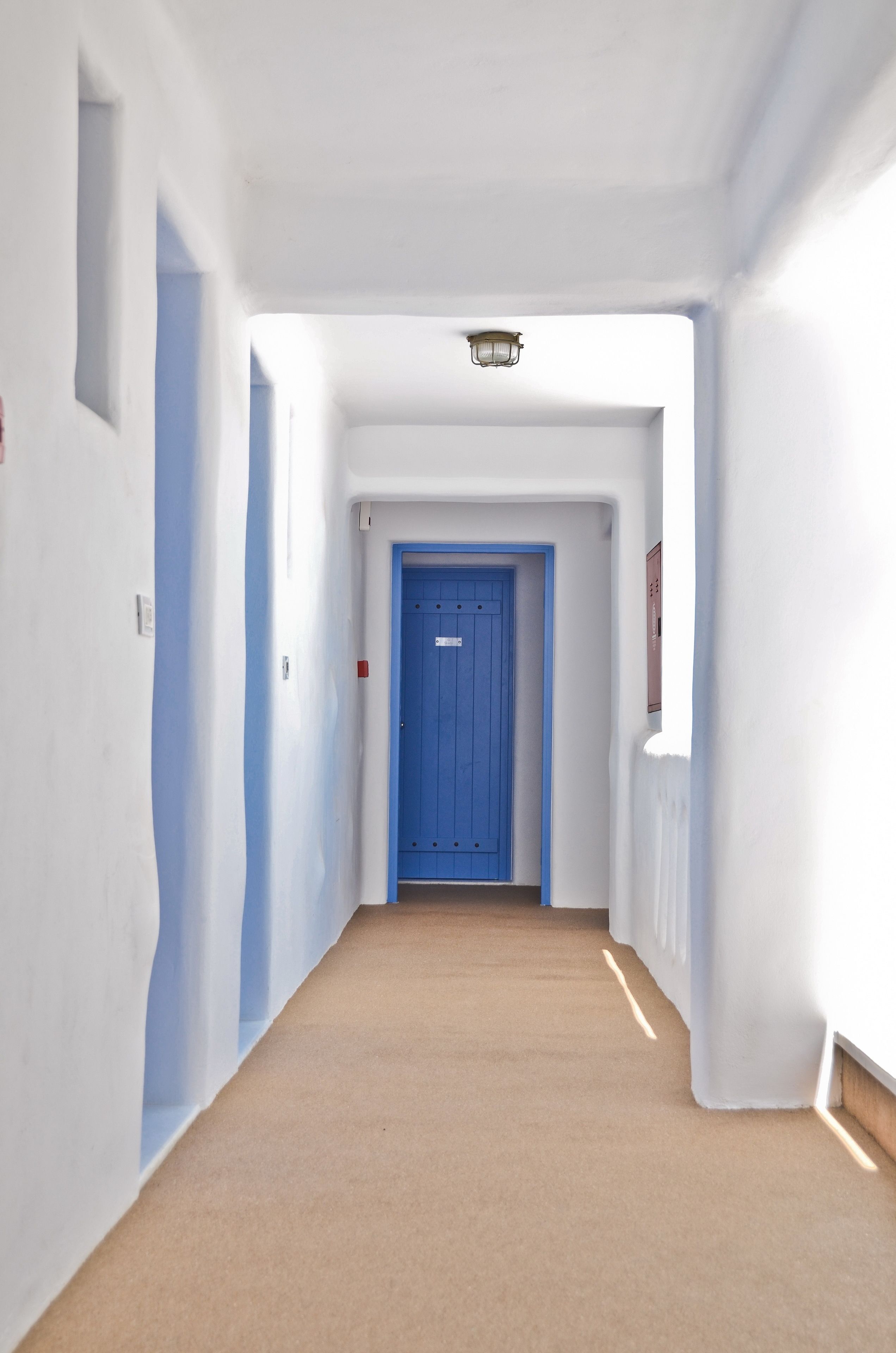 A hallway with a blue door at the end, white walls, and a light fixture above.