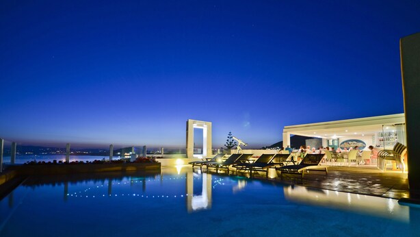 A pool area with lounge chairs and a restaurant, surrounded by a low wall and a view of the cityscape at dusk. - Naxos Island Hotel (Naxos)