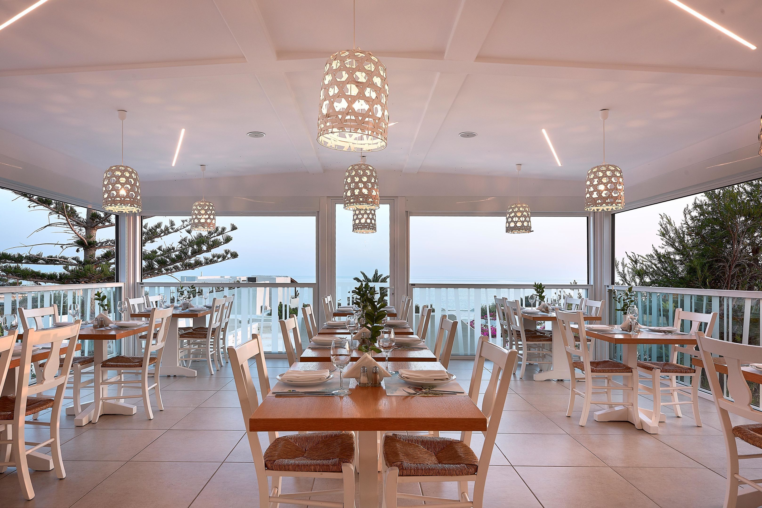 A restaurant with multiple tables and chairs, large windows, and hanging pendant lights.