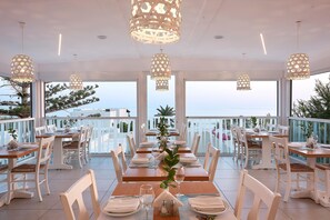 A restaurant with a long dining table set for a meal, surrounded by white chairs, with hanging pendant lights and large windows offering a view of the sea. - Coriva Beach Hotel (Ierapetra)