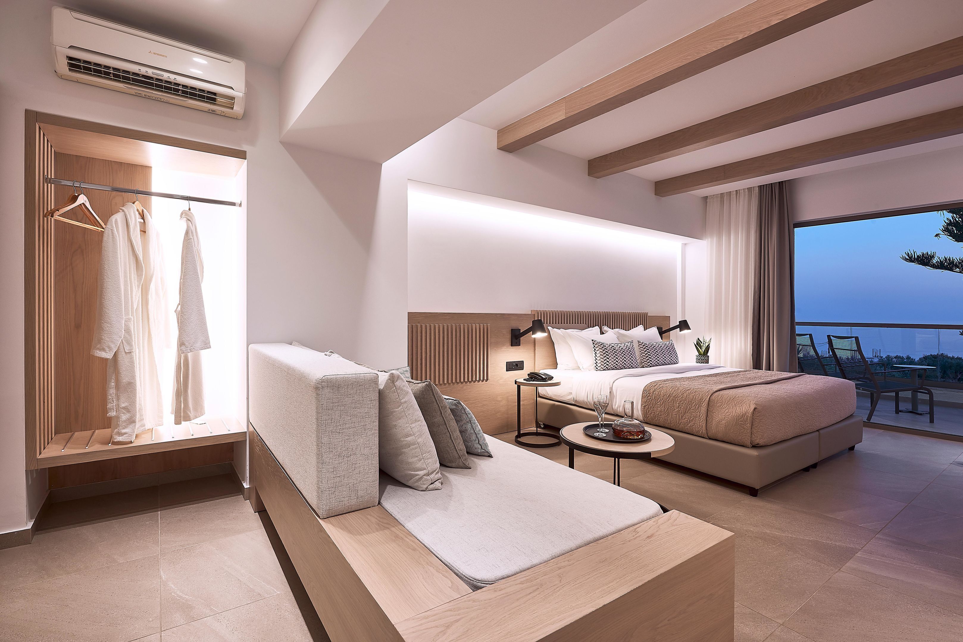 Una habitación de hotel moderna con una cama grande, un sofá, una mesita de noche y un vestidor con batas colgadas.