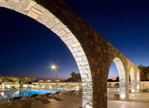 Zona de piscina con arcos de piedra, sillones de descanso y un atardecer de fondo.