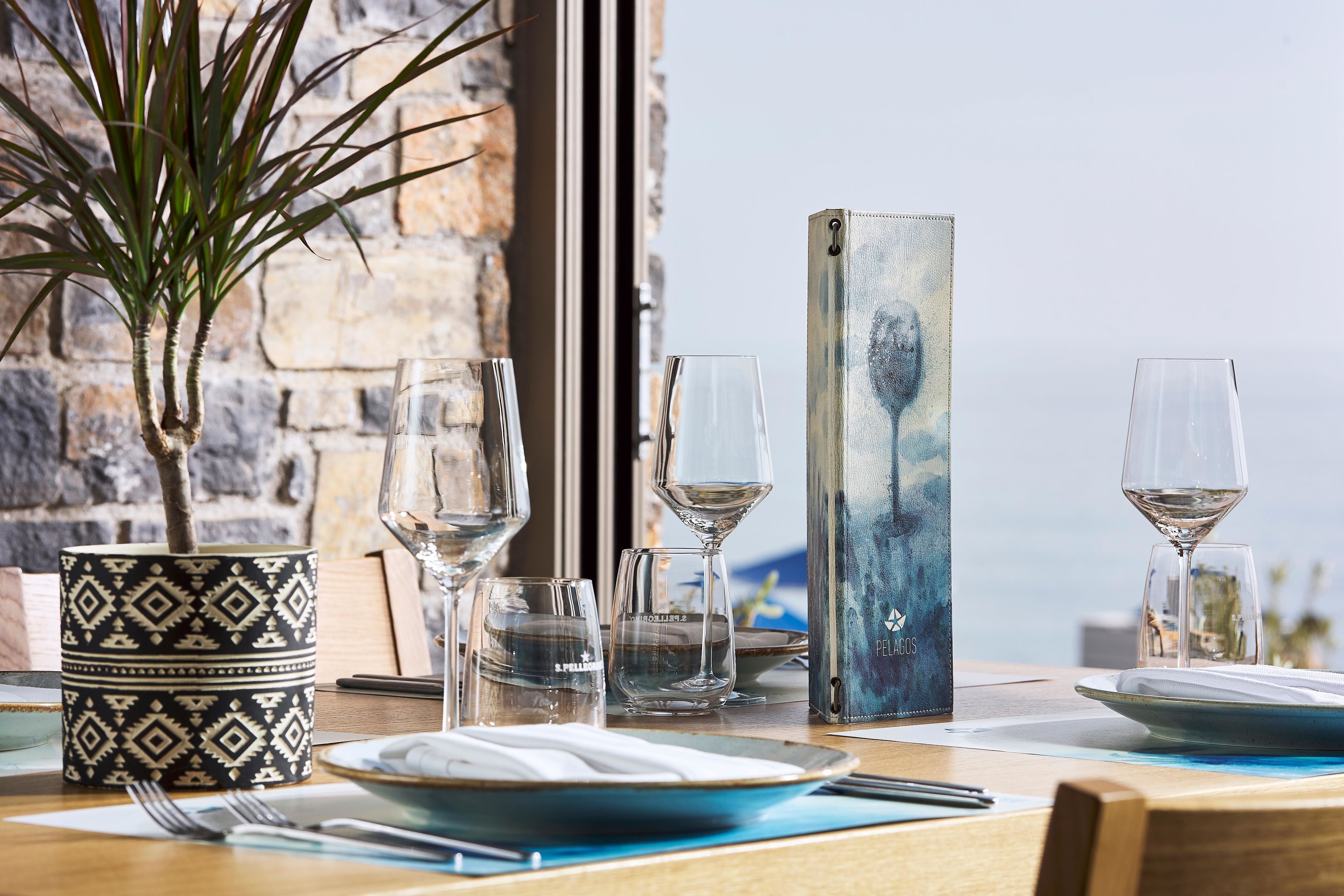 A dining table with plates, glasses, and a menu stand, set against a stone wall with a view.