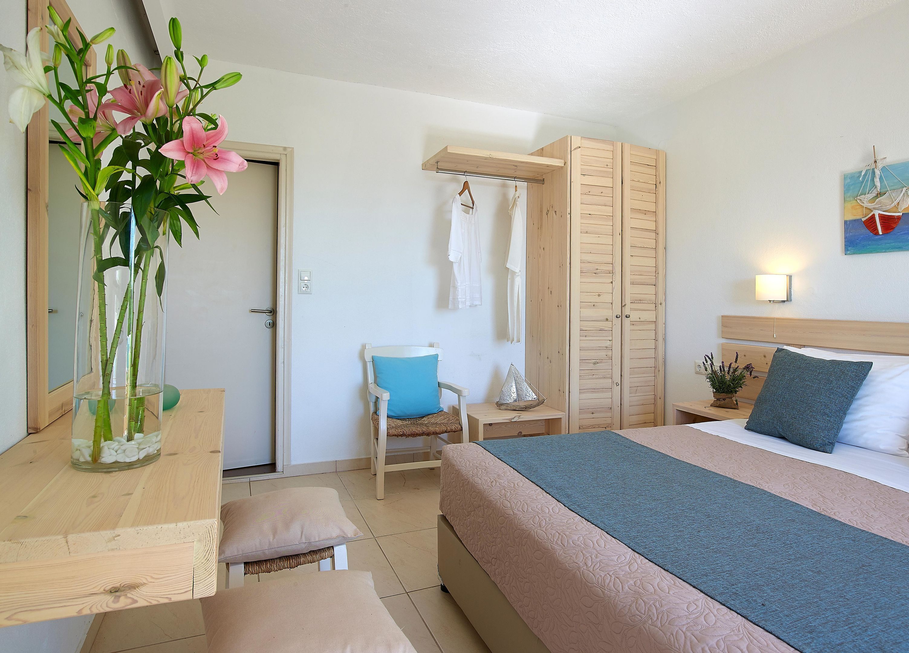 Un dormitorio con cama, una silla, un escritorio, un jarrón con flores, un armario y un cuadro en la pared.