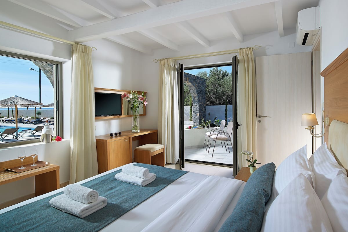 A hotel room with a bed, a desk, a TV, a window with curtains, and a view of a pool and outdoor area.