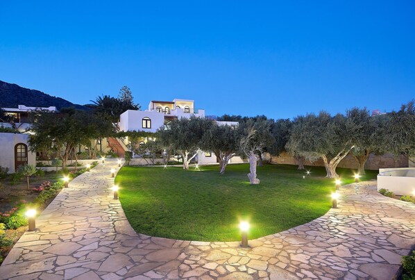 A well-lit courtyard with a stone pathway, surrounded by trees and a building in the background. - Coriva Beach Hotel (Ierapetra)