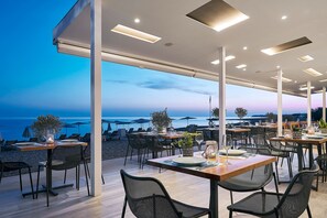 An outdoor dining area with tables and chairs, overlooking a beach and the sea. - Coriva Beach Hotel (Ierapetra)