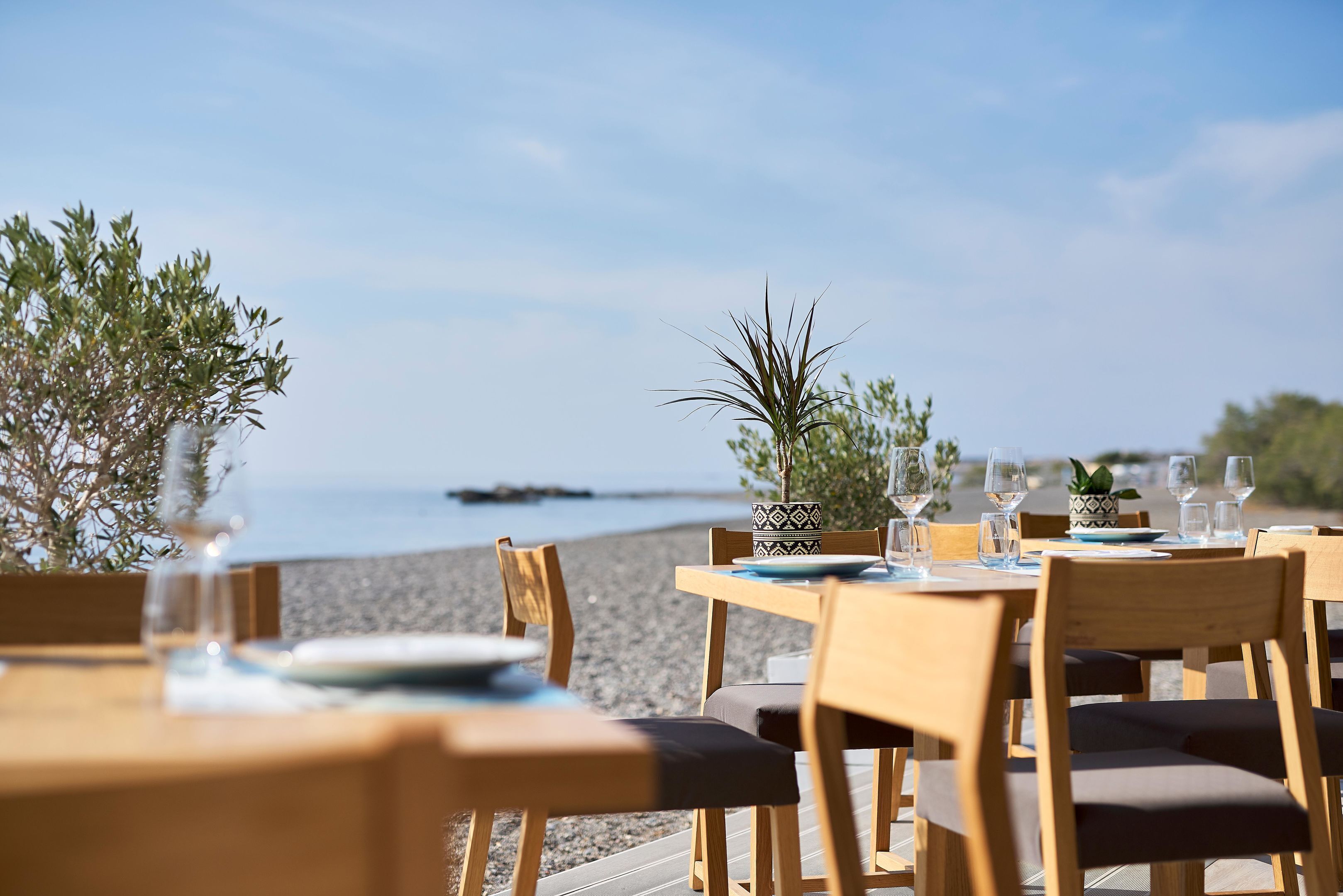 A seaside restaurant with wooden tables and chairs, a view of the beach, and a calm sea in the background.