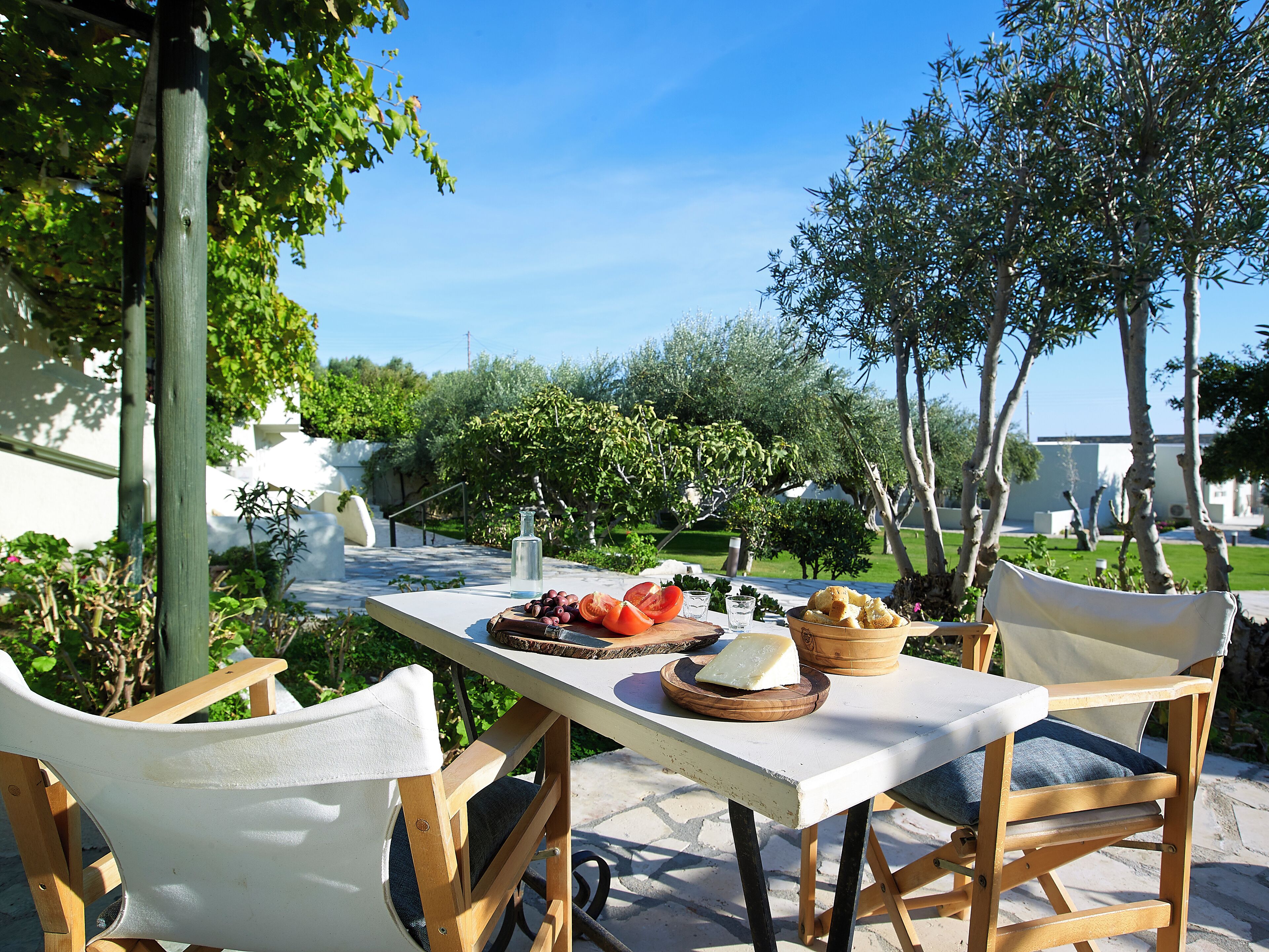 A table set outdoors with a variety of food items, including cheese, olives, and bread, surrounded by wooden chairs.