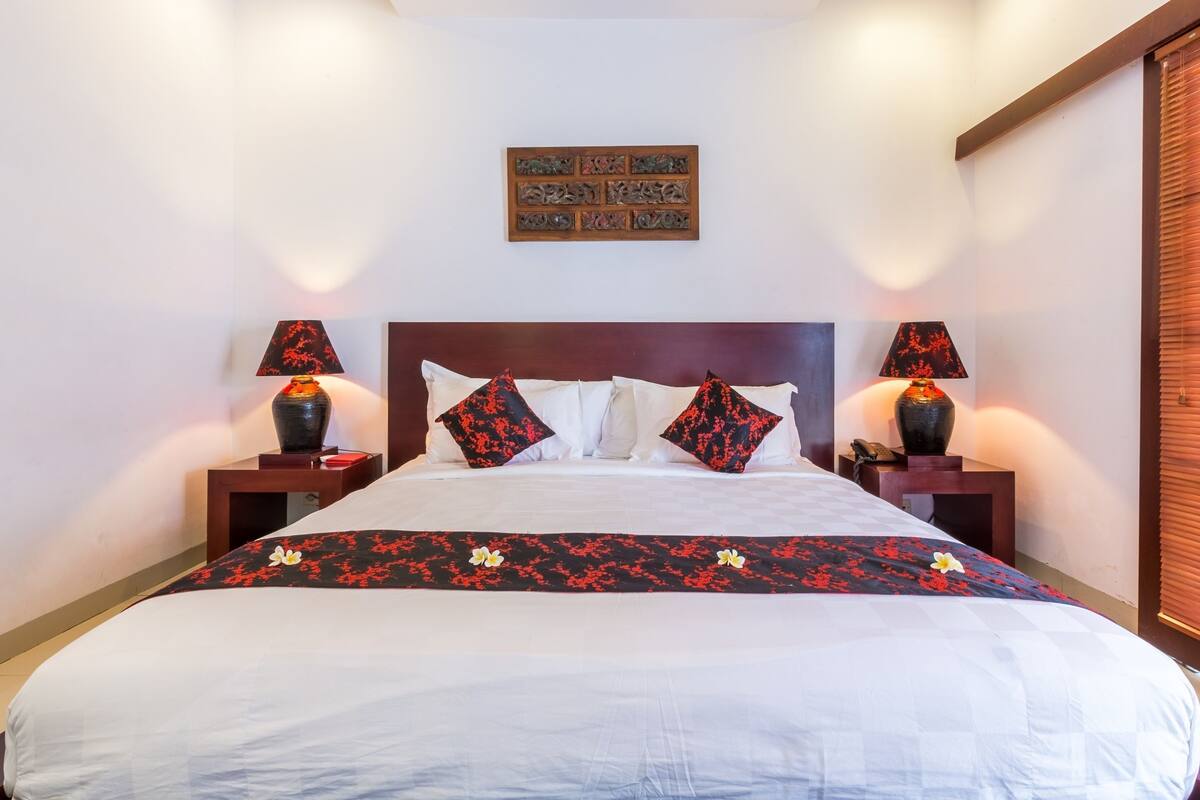 Una cama bien hecha con sábanas blancas y almohadas estampadas con flores rojas, dos mesitas de noche con lámparas y un cabecero de madera.