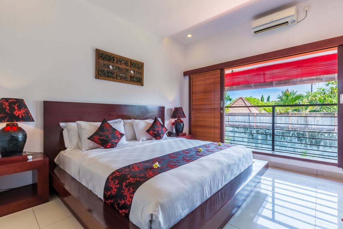 Habitación de hotel con una cama grande, cabecera de madera, dos mesitas de noche con lámparas, un adorno en la pared y un balcón con toldo rojo.