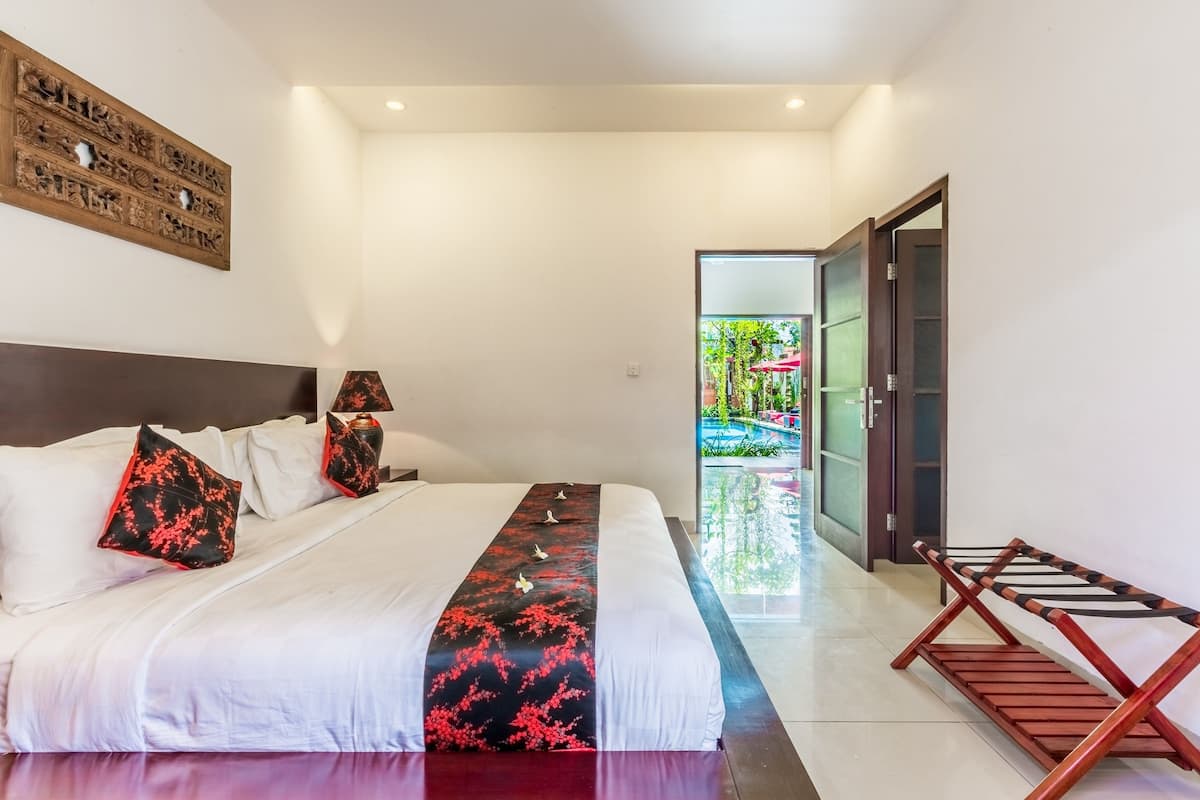 Habitación de hotel moderna con una cama grande, cabecera de madera, dos mesitas de noche con lámparas y vistas a una piscina y follaje a través de una puerta abierta.