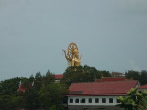 A large golden statue atop a hill with a red-roofed building in the foreground. - The Lounge - Hostel (Koh Samui)