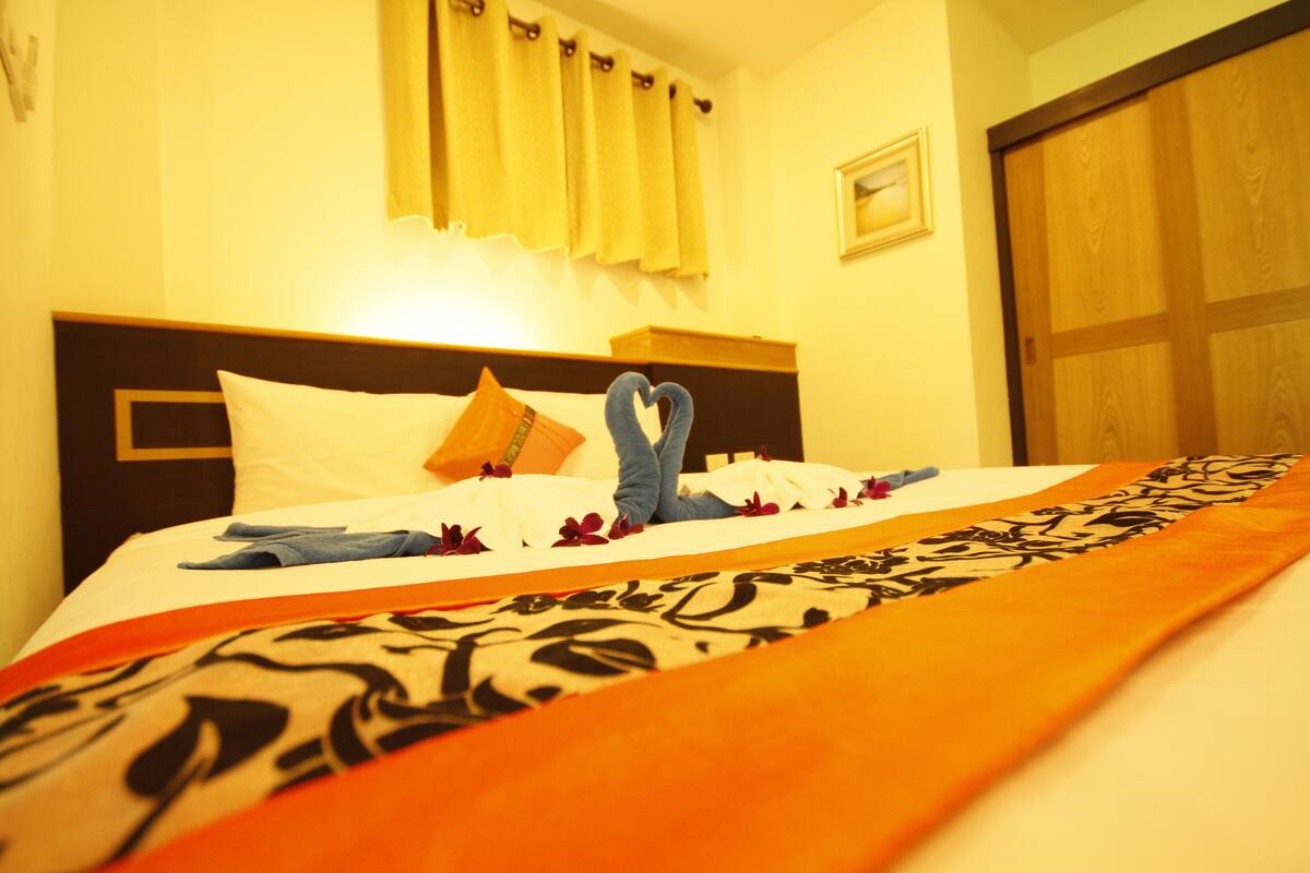 A neatly made bed with a patterned bedspread, a pillow, and a heart-shaped towel arrangement.
