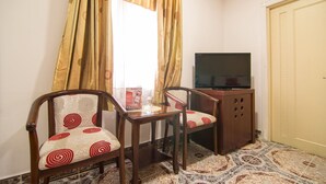 Quarto pequeno com duas cadeiras de madeira, uma mesa de vidro, televisão sobre um móvel de madeira e um tapete estampado.