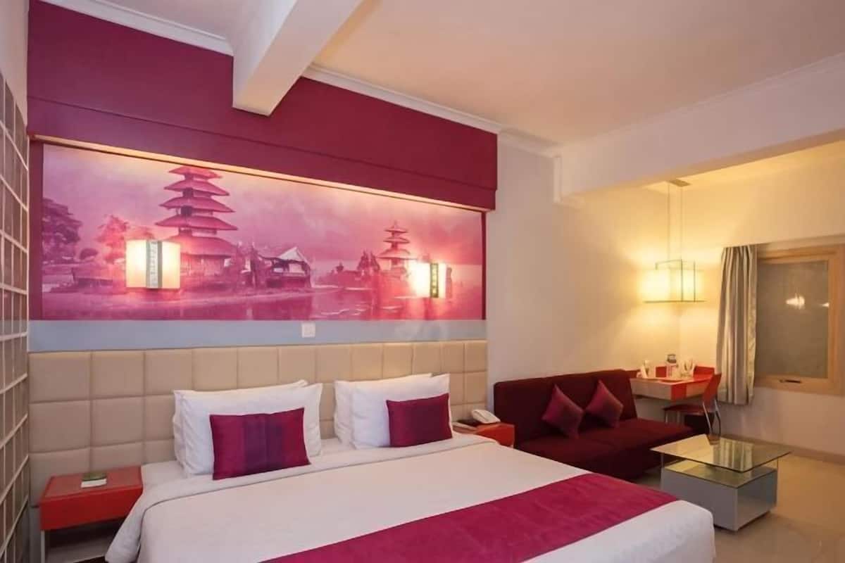 A hotel room with a bed, a red sofa, a glass coffee table, a nightstand, and a wall-mounted lamp.