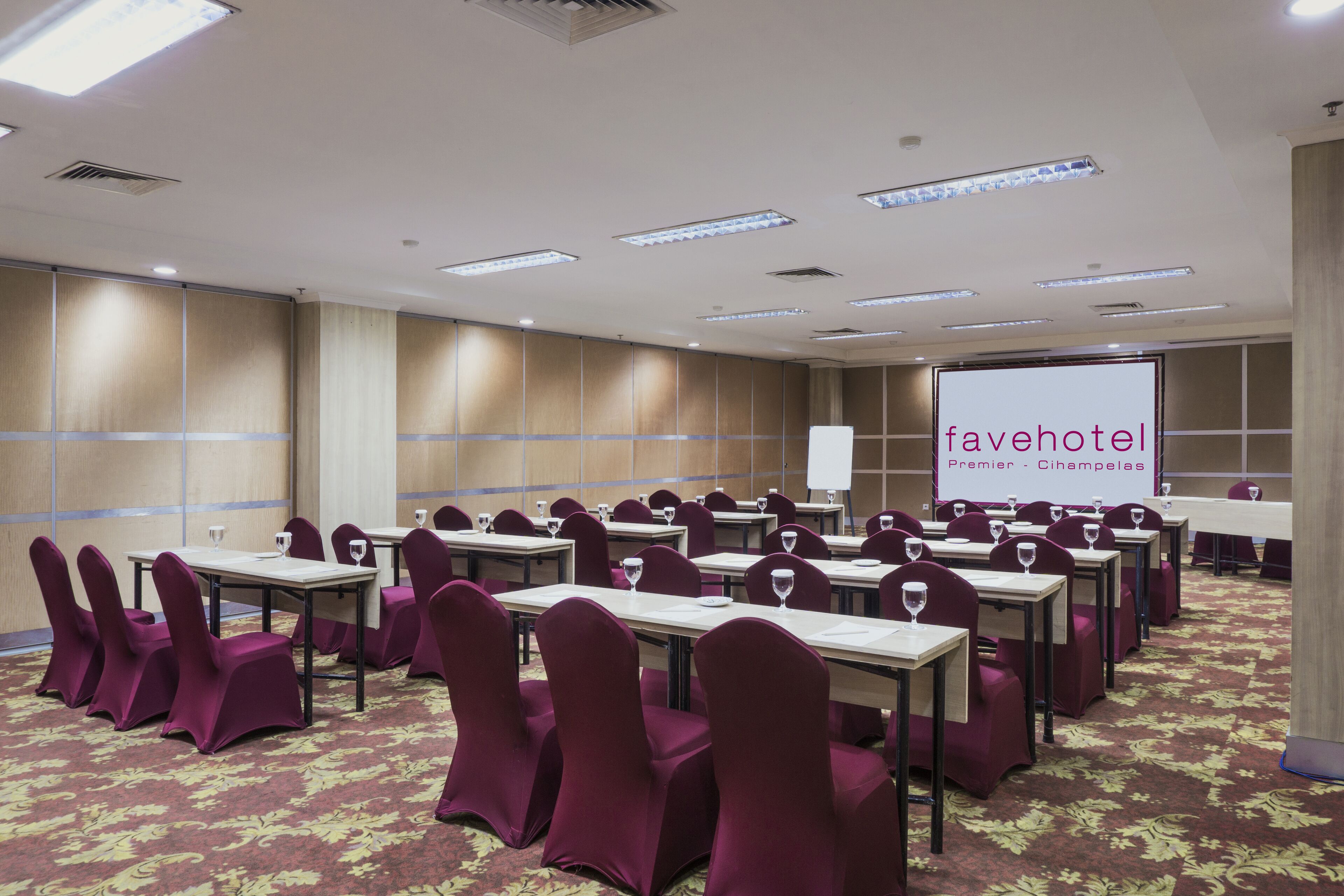 Un salón de conferencias con filas de mesas y sillas, una pantalla grande mostrando 'fayehotel Premier - Cihampelas' y un suelo alfombrado.