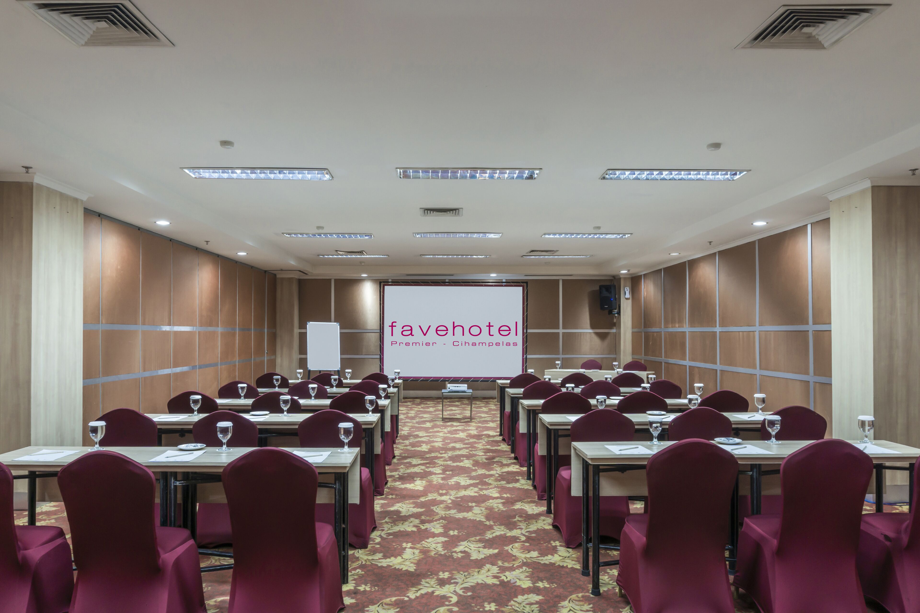 A conference room with rows of tables and chairs, a large screen at the front, and a carpeted floor.