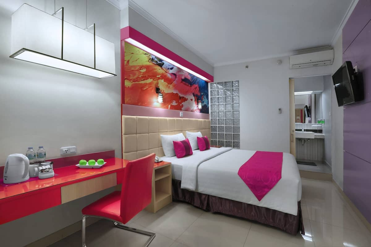 A modern hotel room with a bed, a desk, a chair, a TV, and a bathroom visible through an open door.