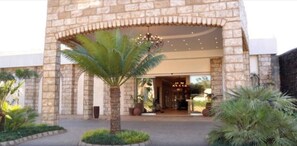 Un edificio de piedra con una palmera grande en el centro, un acceso cubierto y un candelabro visible en el interior.
