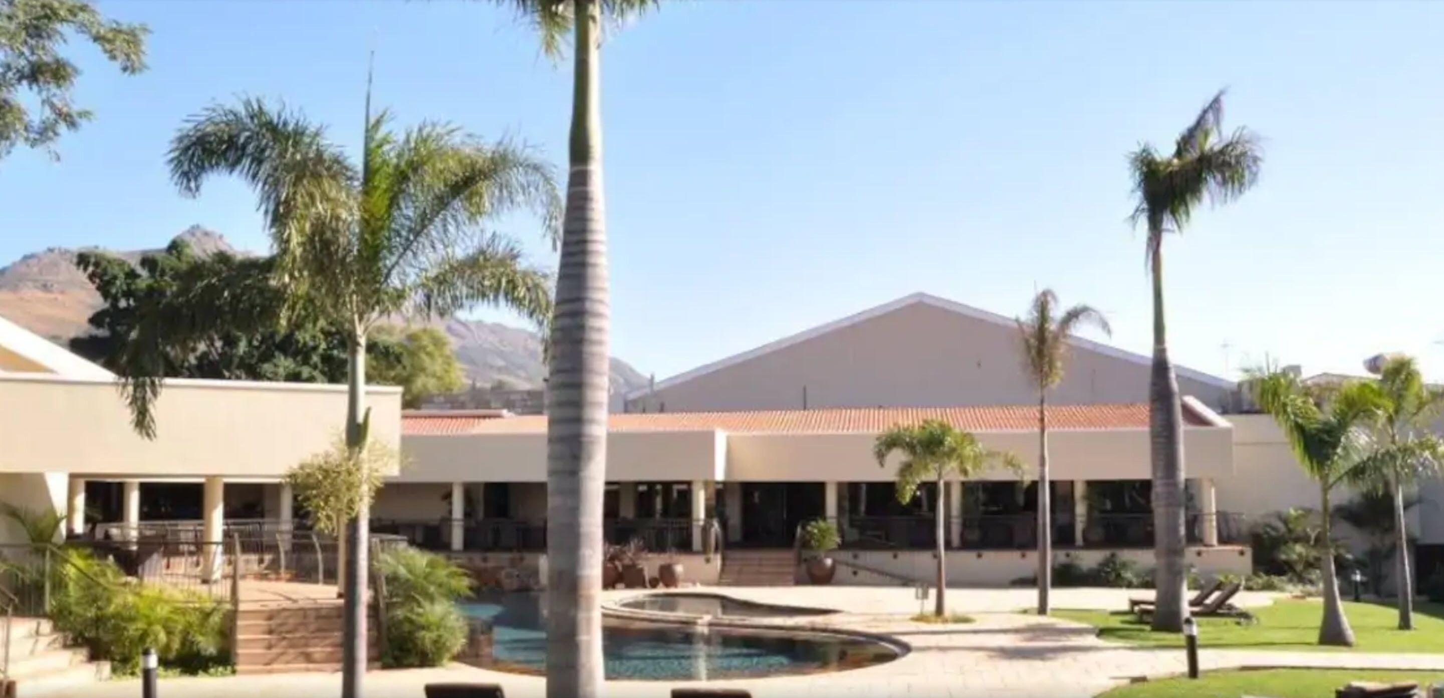 A resort with a swimming pool, palm trees, and a mountain backdrop.
