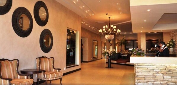 Vestíbulo de hotel con mostrador de recepción, área de descanso con sillones, candelabro y pared con espejos circulares.