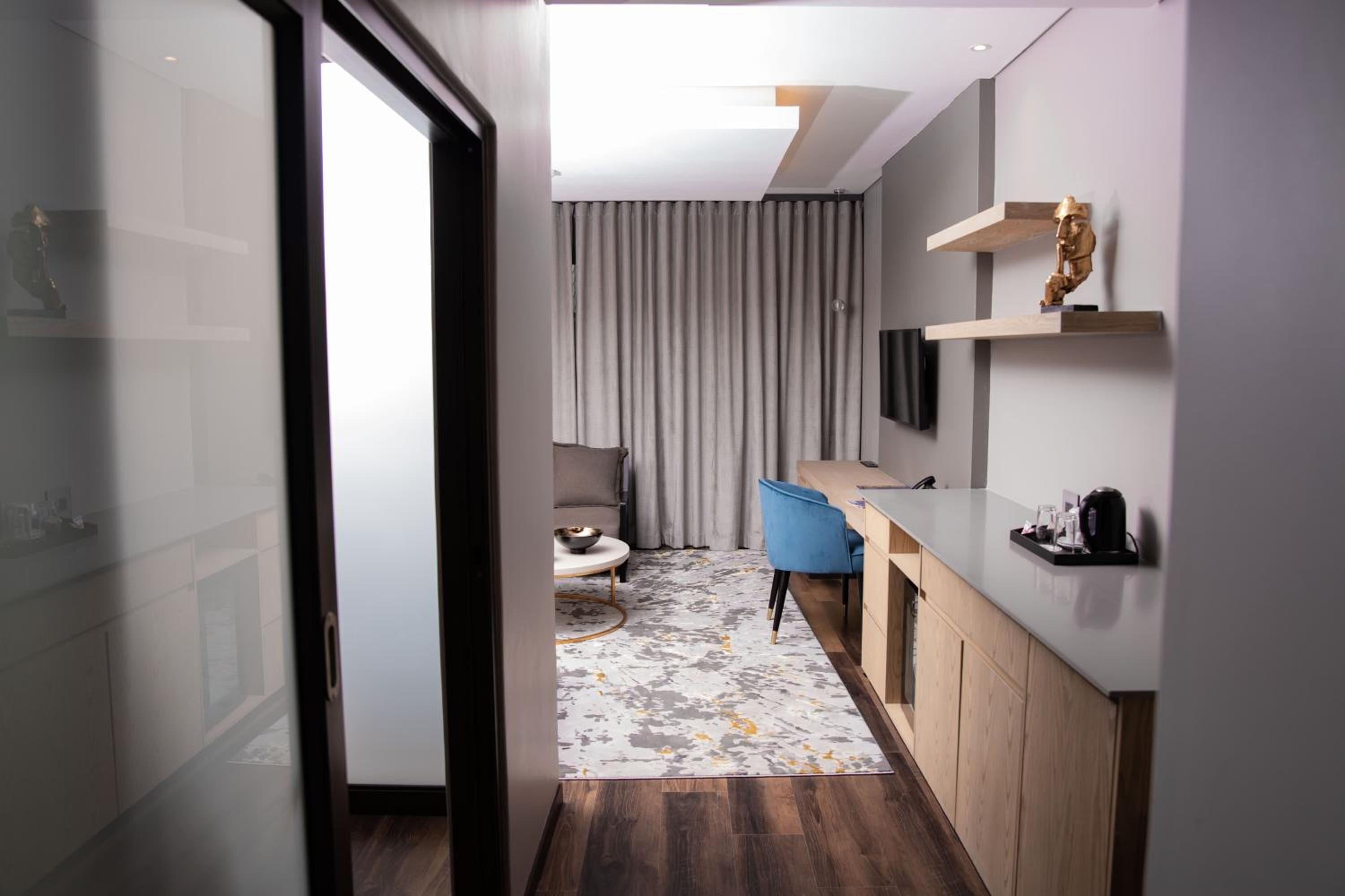 A modern hotel room with a glass door leading to a balcony, a small kitchenette, a seating area with a sofa and coffee table, and a large window with curtains.