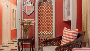 Una habitación con suelo a cuadros, un sofá de madera con un cojín a rayas, una pequeña mesa con un jarrón de flores y un espejo decorativo en la pared.
