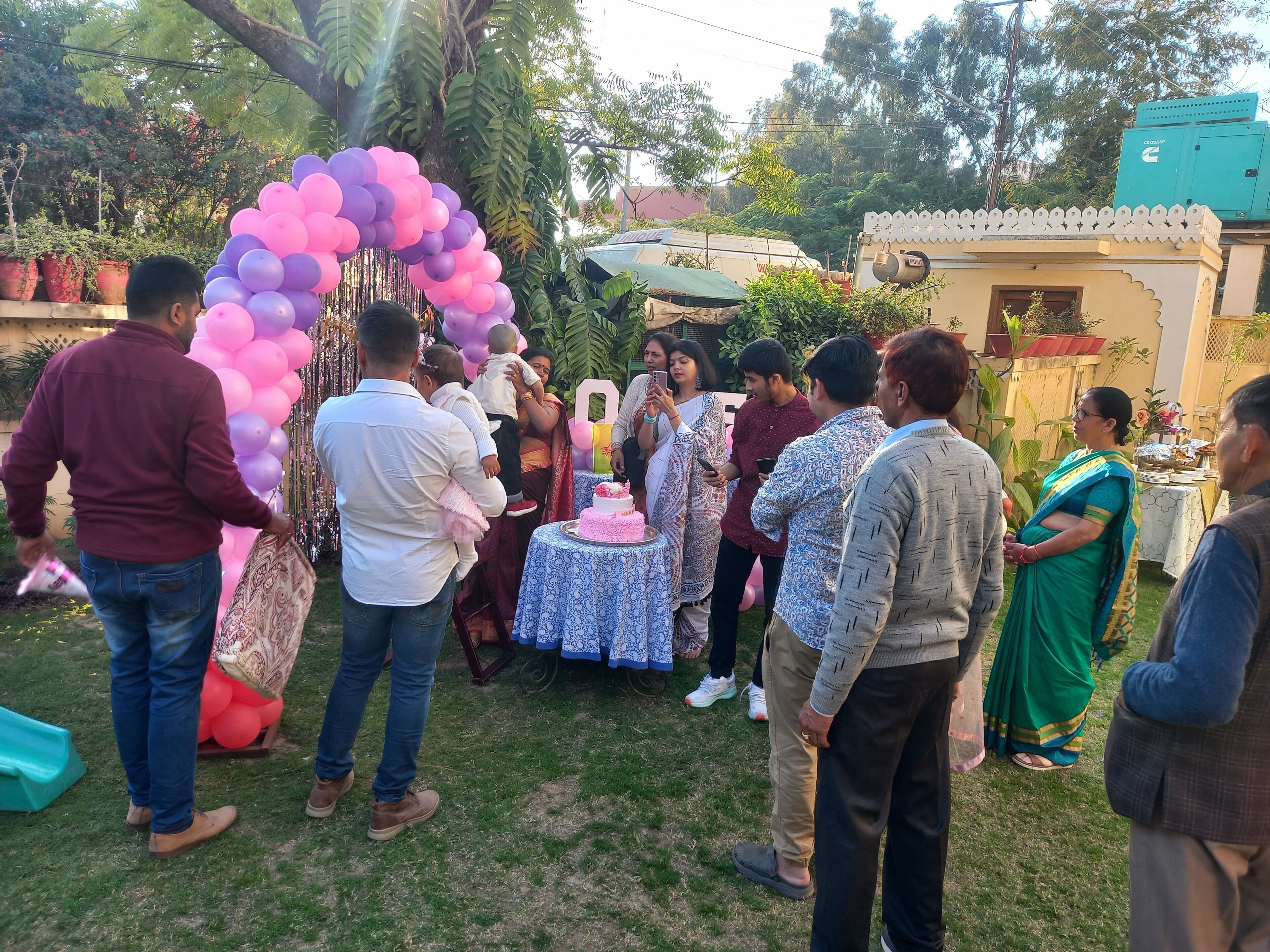A group of people gathered around a decorated area with a table and a cake, suggesting a social event.