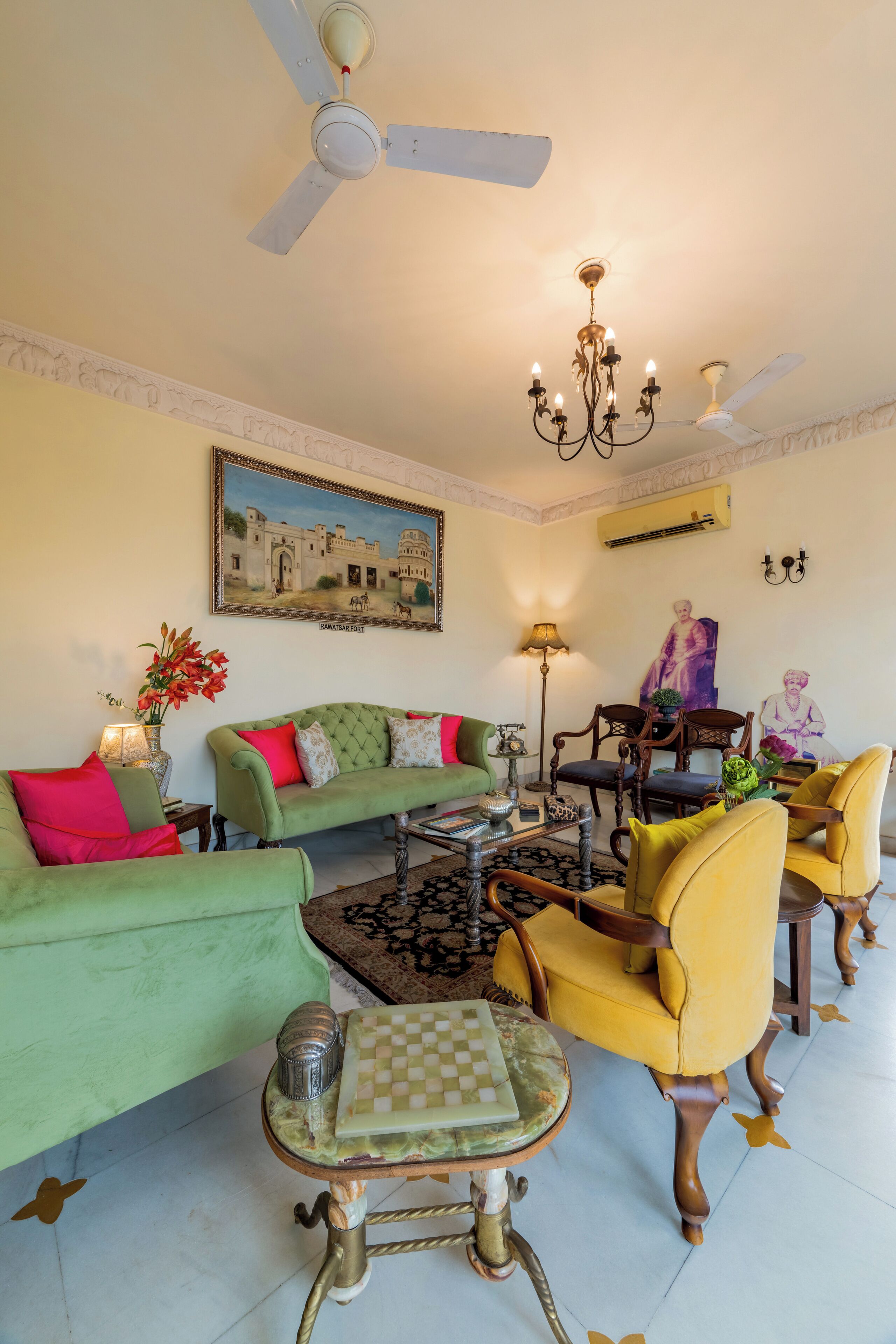 Una sala de estar con un sofá verde, sillones amarillos, una araña, ventiladores de techo, un tablero de ajedrez y un cuadro de un edificio.